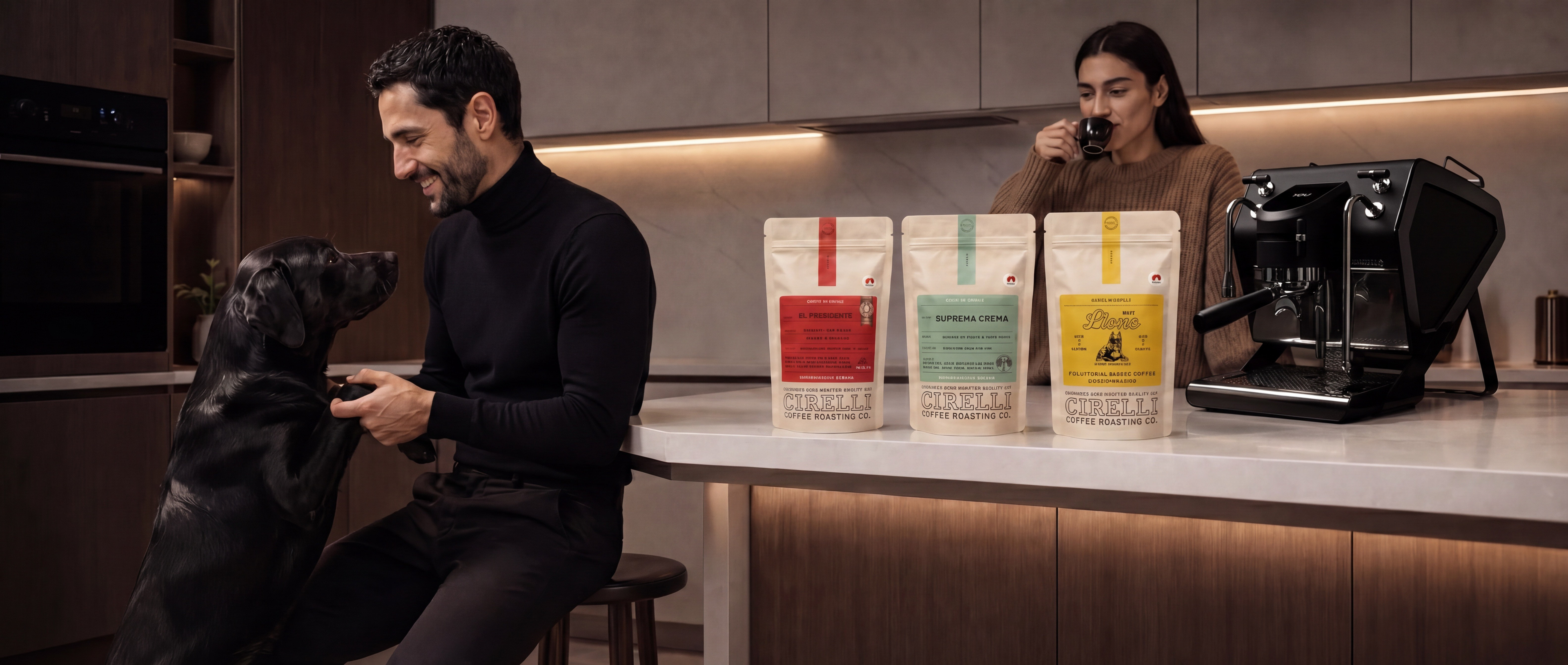 A man in a black turtleneck sits on a stool in a modern kitchen, petting a black dog. A woman in a brown sweater stands behind the counter drinking from a mug, with three standing bags of dog food and a black espresso machine on the counter.