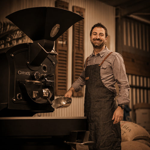 A smiling man in a gray shirt and dark denim apron stands beside a large black Giesen coffee roaster in a rustic workshop setting with warm sepia lighting.