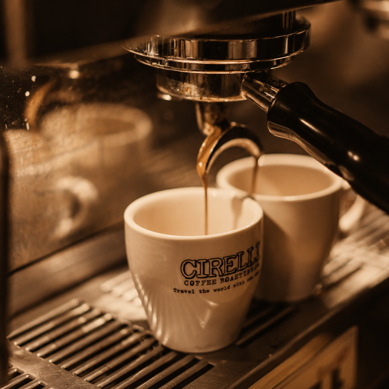 A close-up shot of espresso being poured from an automatic coffee machine into two white ceramic cups, one of which has the brand name 'CIRELLI COFFEE ROASTERS' printed on it.