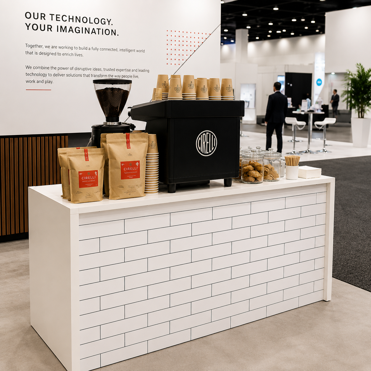 A white brick-patterned coffee station counter with a black espresso machine, paper cups, coffee bags, and a coffee maker, set in a large exhibition hall with a white wall in the background.