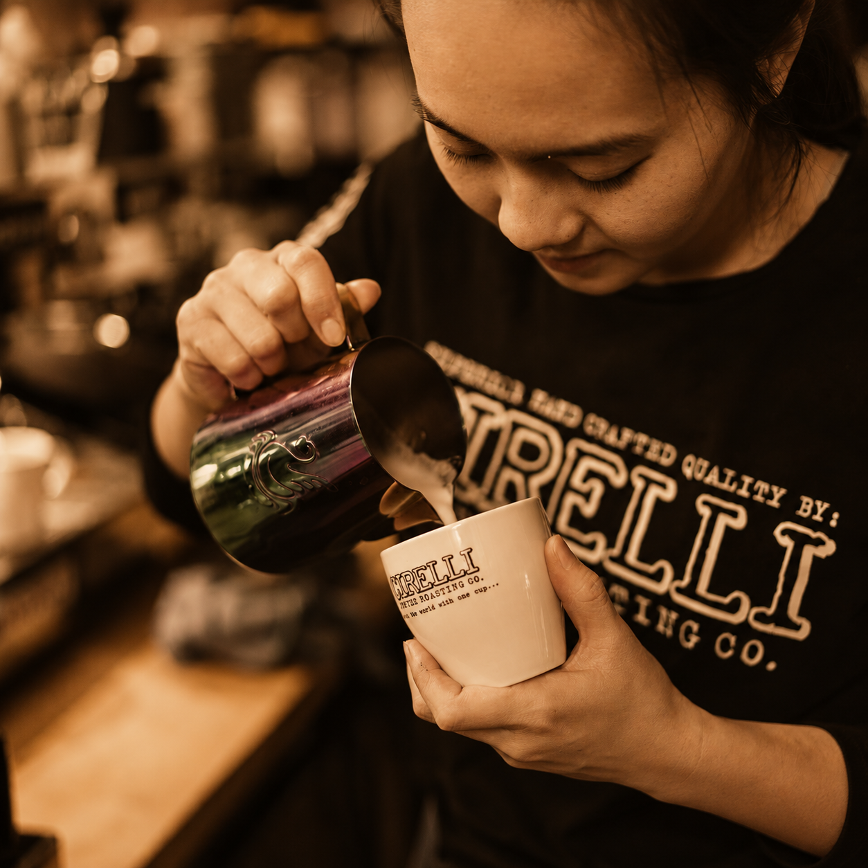 A barista pouring milk from a metallic pitcher into a white ceramic cup printed with the brand name 'Trelli'. The person is wearing a black T-shirt with 'CIRELLI' printed in beige, and the background is a warm, blurred coffee shop setting.
