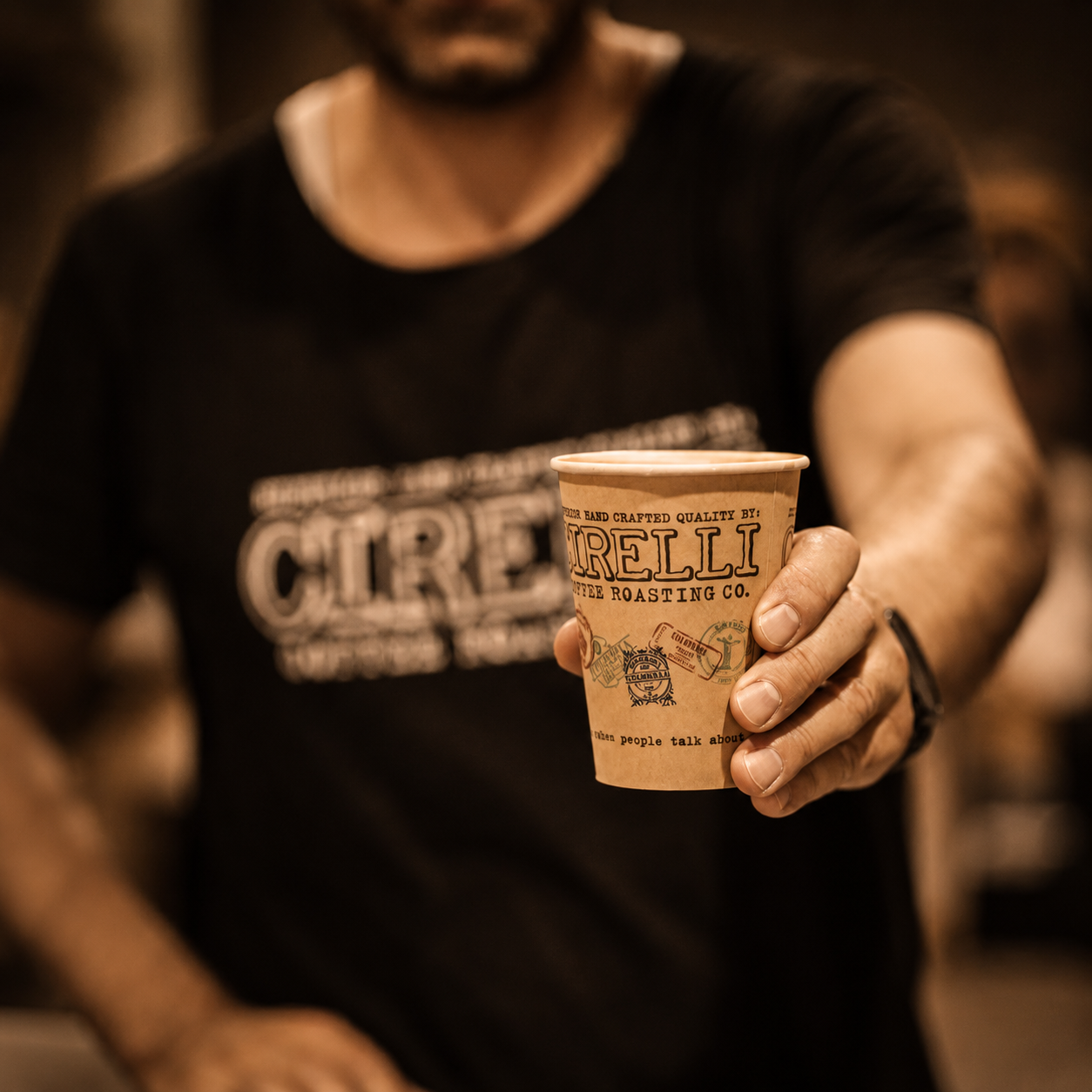 A person in a black graphic T-shirt holds up a small kraft paper coffee cup printed with the brand name 'Cattle Roasting Co.' The background is heavily blurred with warm sepia tones, and the person is out of focus except for the torso and hand holding the cup.