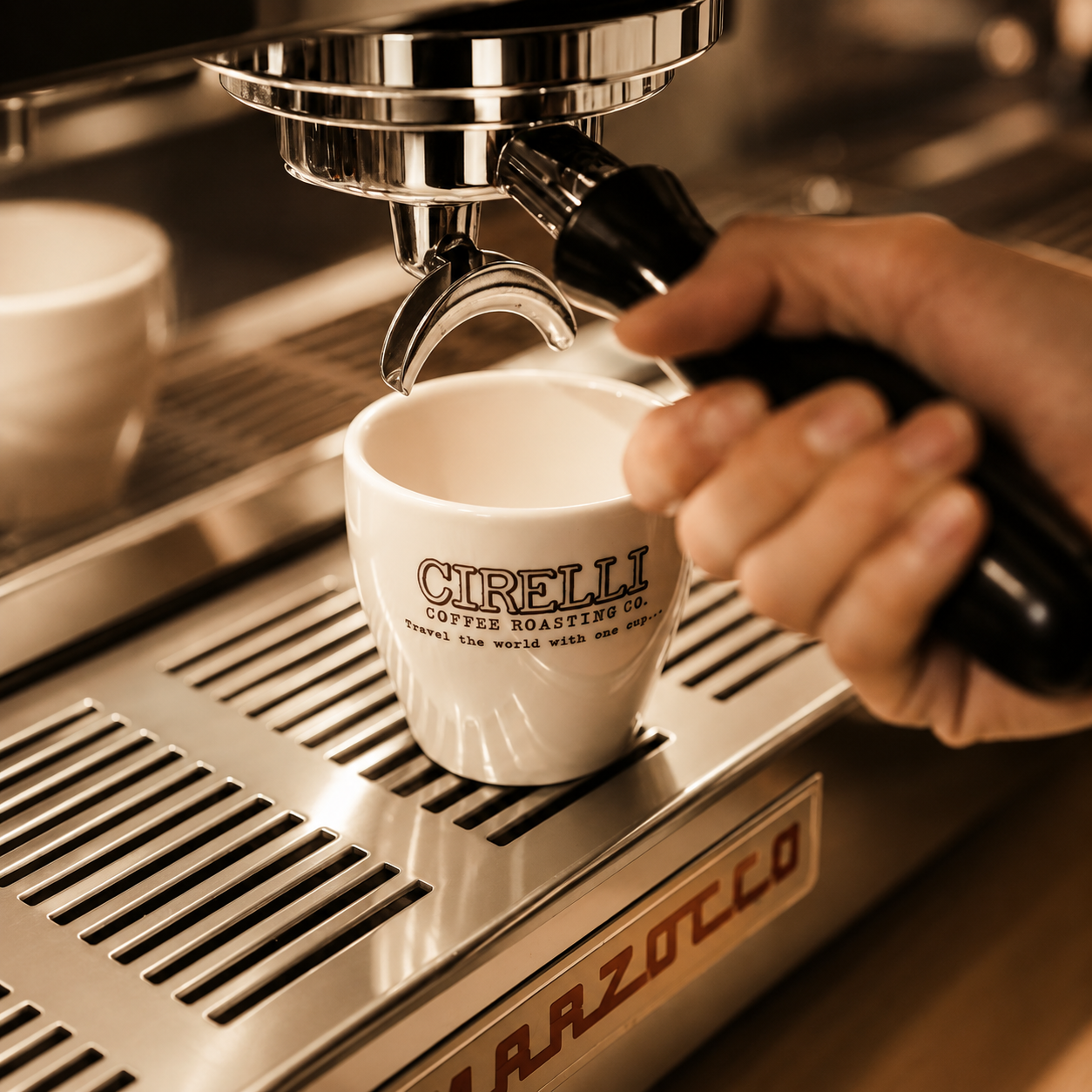 A close-up of a hand pressing a black handle to dispense espresso into a small white Cirelli coffee cup on a stainless steel espresso machine.