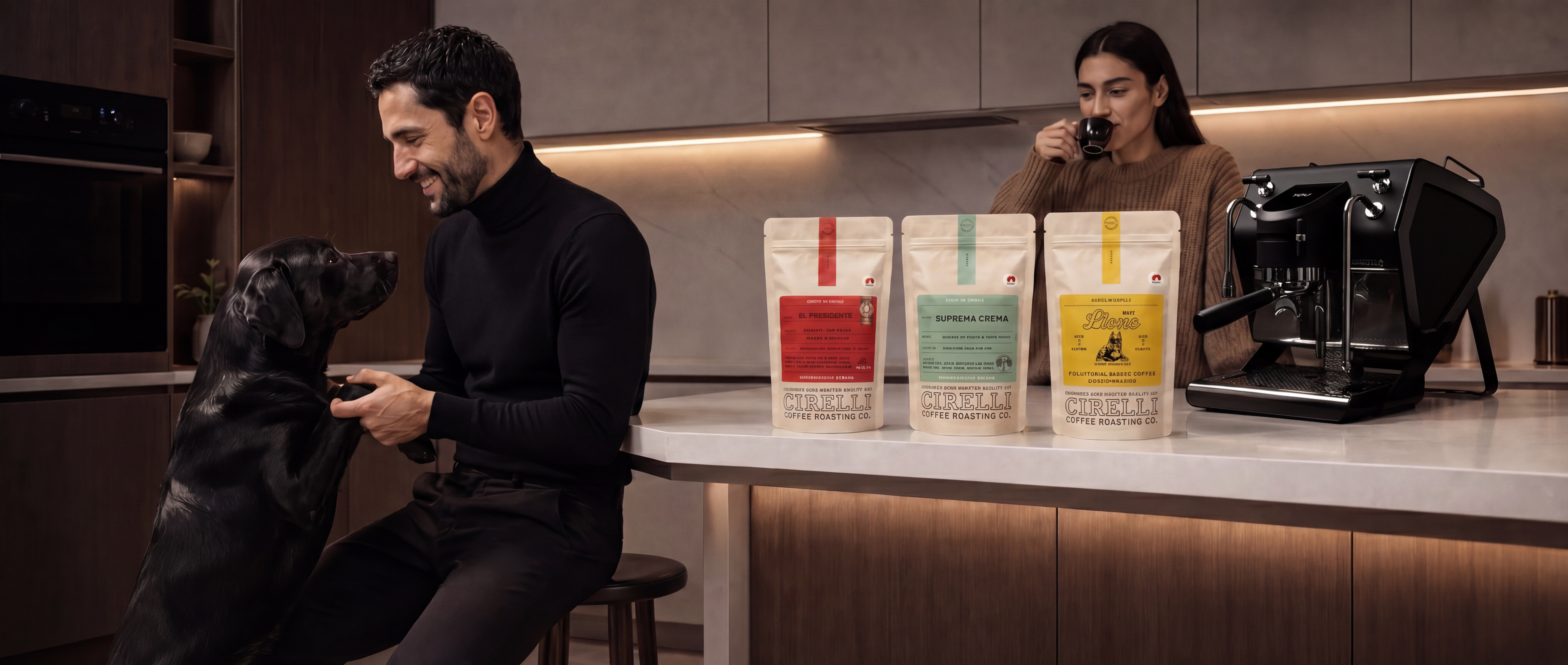 A man in a black turtleneck sits on a stool in a modern kitchen, petting a black dog. A woman in a brown sweater stands behind the counter drinking from a mug, with three standing bags of dog food and a black espresso machine on the counter.