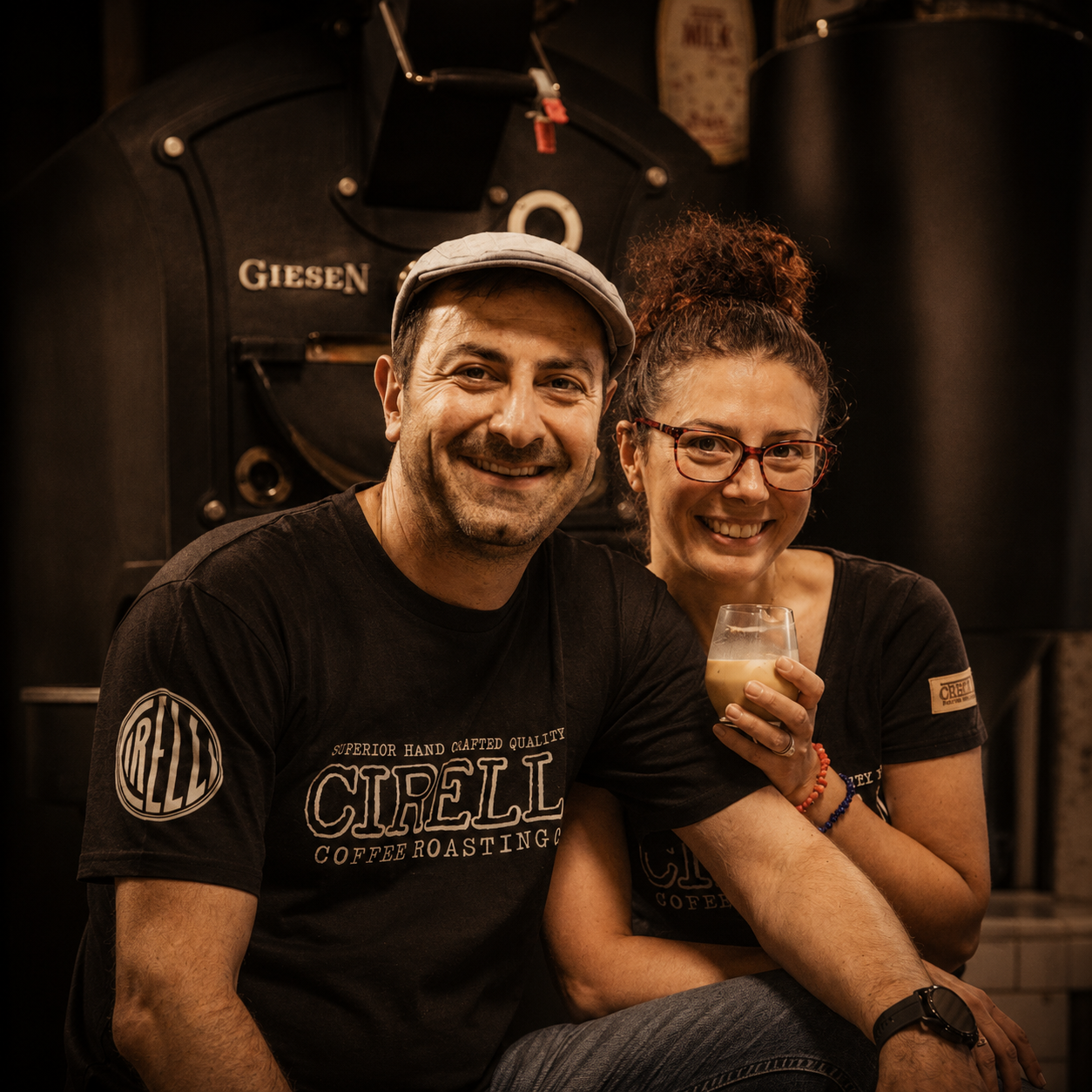 Two people smiling in front of a coffee roaster, wearing matching black Ciellet coffee roasting t-shirts. The man wears a light-colored cap, and the woman holds a glass of coffee.