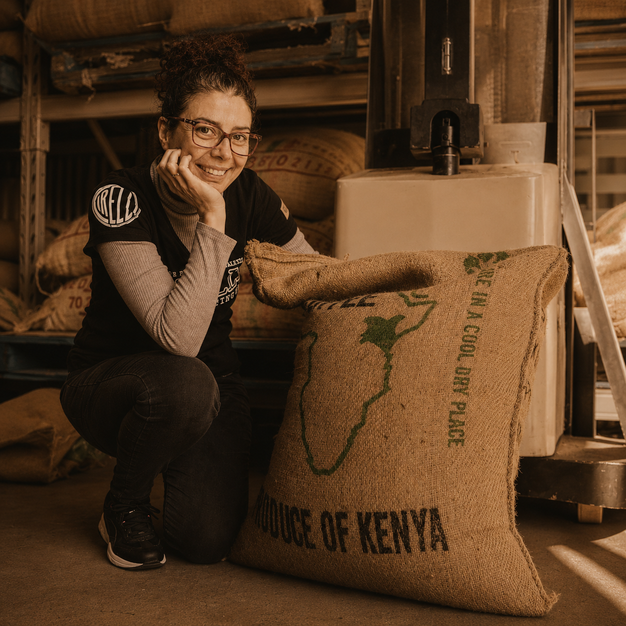 A woman wearing glasses and a black-and-gray shirt crouches beside a large burlap coffee sack printed with 'PRODUCE OF KENYA' and a green map outline, in a warm, rustic warehouse setting.