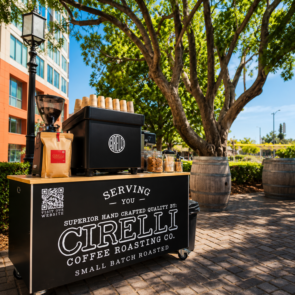 A black Cirelli coffee roasting cart set outdoors on a brick walkway under a large tree, with coffee equipment, cups, and branded signage visible.