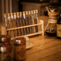 A wooden rack holding multiple clear glass test tubes filled with dark roasted coffee beans, set on a warm wooden table with a coffee maker and bottle in the background.