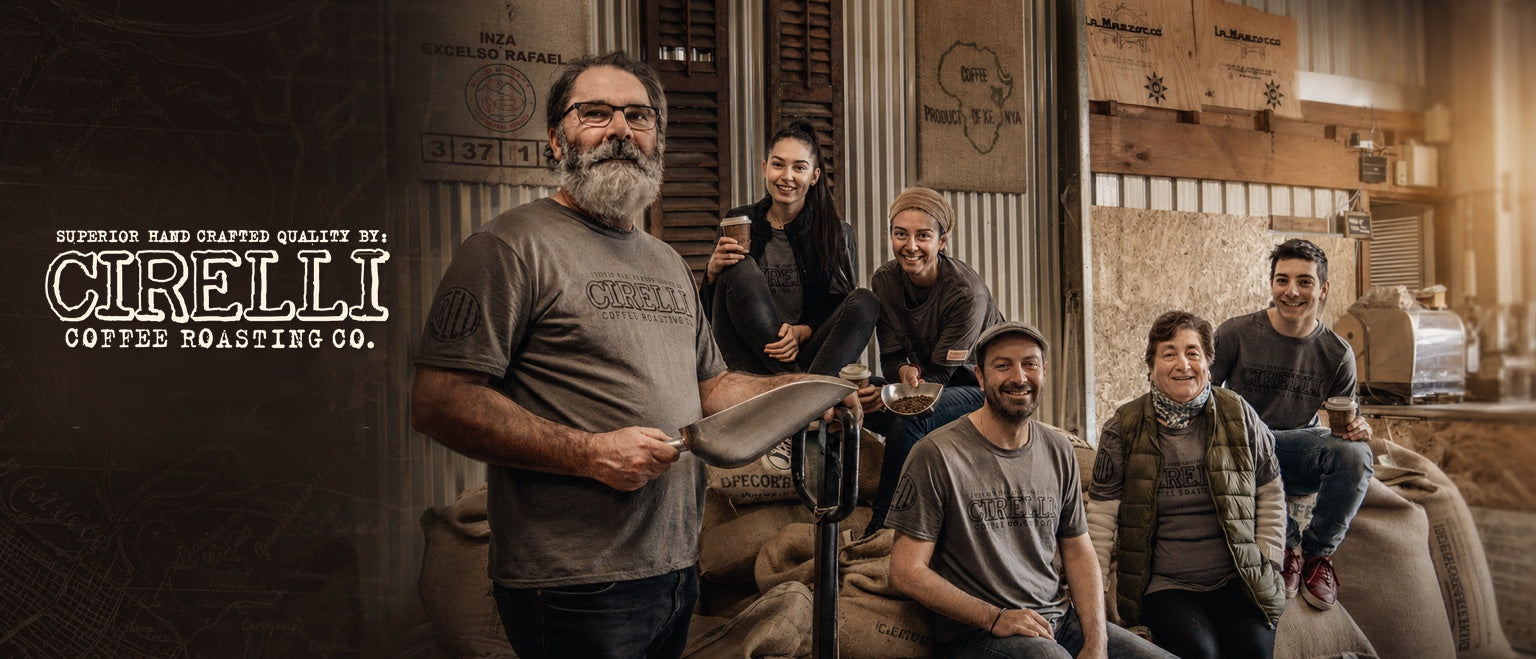 A group of six people posing together in a rustic coffee roasting facility, wearing matching gray t-shirts with the CIRELLI logo. The background features wooden walls, cardboard boxes, and a large coffee roaster.